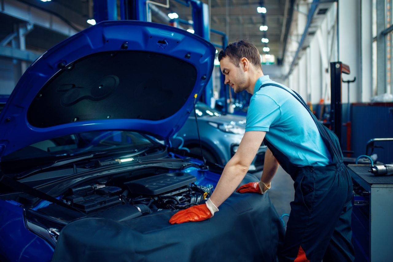 Mechanic in overalls working under the bonnet of a blue car at a car workshop
